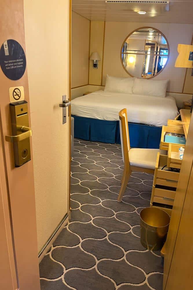 Cruise ship room with neatly made bed, round mirror above headboard, desk and chair, patterned carpet, and open door with no-smoking sign.