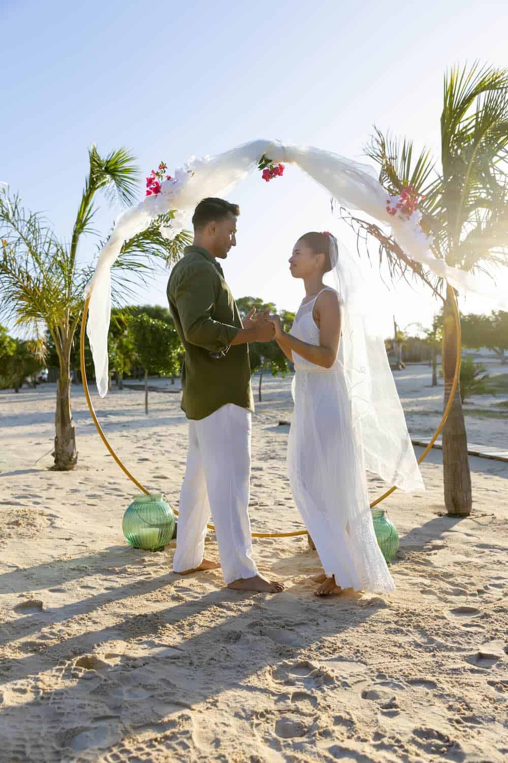 A couple stands under a decorated arch on a beach, holding hands and facing each other, with palm trees and sunlight in the background.