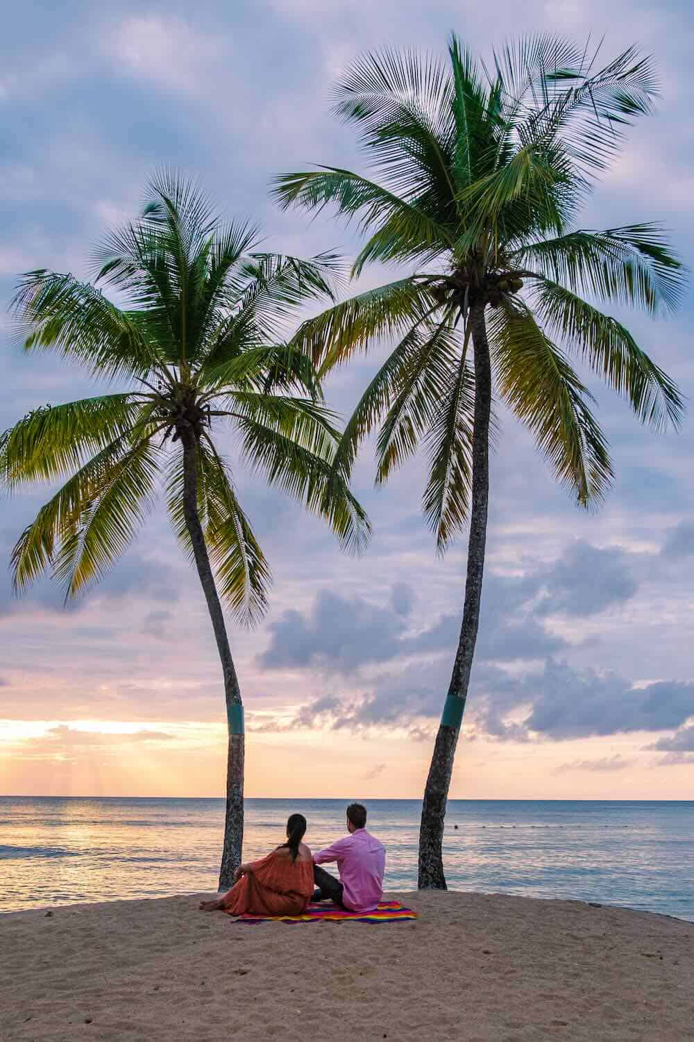Two people sit on a blanket on a sandy St. Lucia beach at sunset, facing the ocean, with two tall palm trees framing the scene.