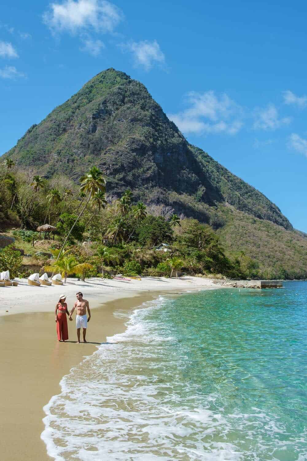 A couple walks along a beach in St. Lucia with clear turquoise water, palm trees, and a tall green mountain in the background under a blue sky.