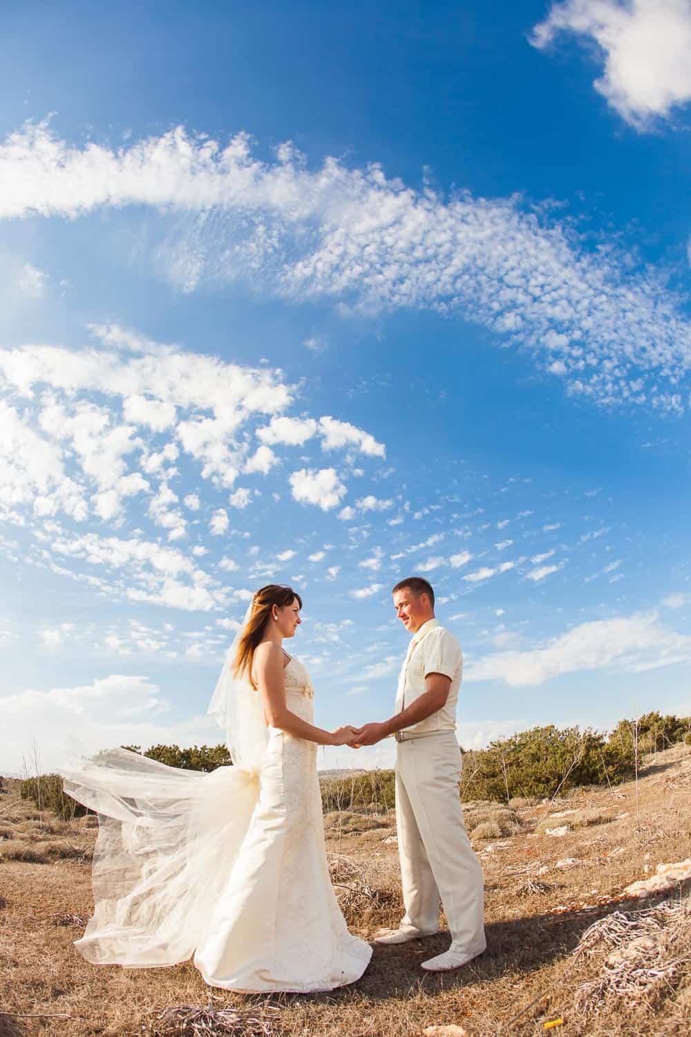 A bride and groom stand outdoors holding hands, facing each other, with a bright blue sky and wispy clouds in the background.