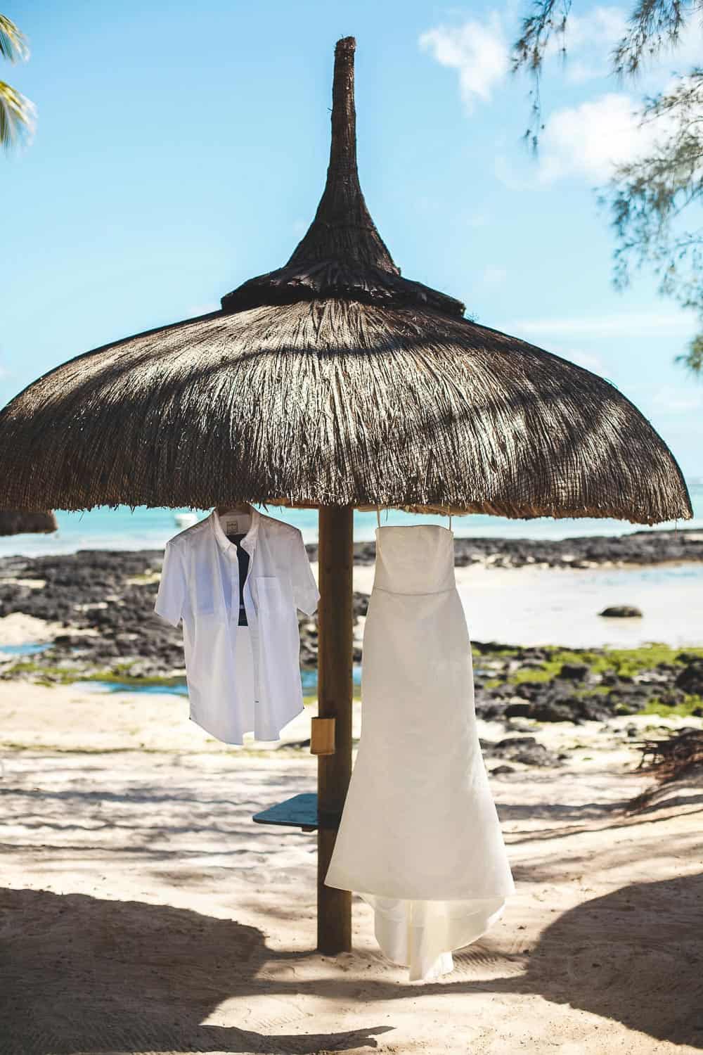 A white dress and a white shirt hang from a thatched beach umbrella, with sand, rocks, and the ocean in the background.