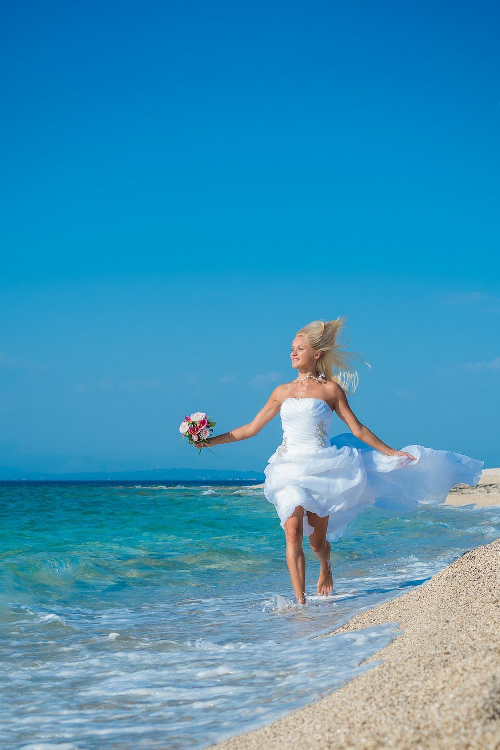 A woman in a white wedding dress holding a bouquet walks along the shoreline with waves at her feet under a clear blue sky.
