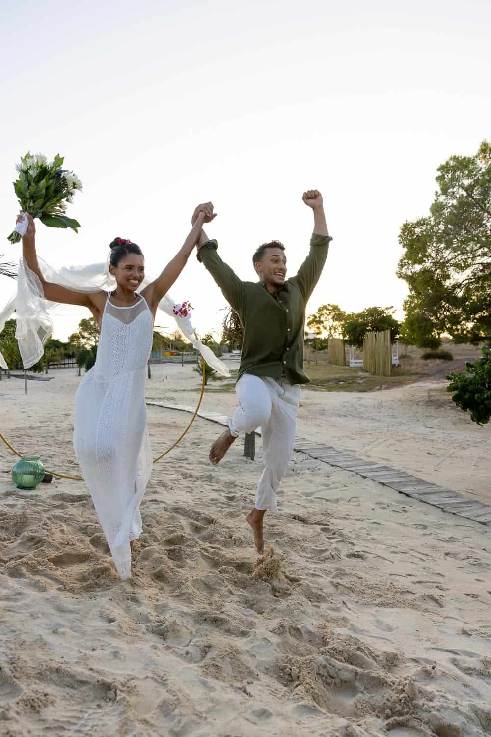 A couple, holding hands and smiling, run barefoot on a beach; the woman wears a white dress and holds a bouquet, while the man raises his arm in celebration.