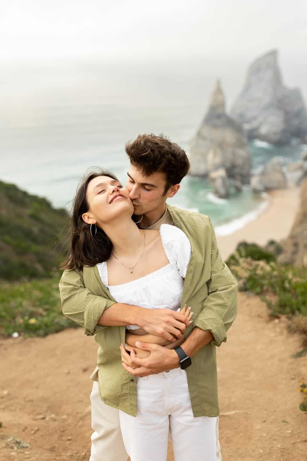 A man embraces and kisses a woman on the cheek as they stand on a coastal path with cliffs and the sea in the background.