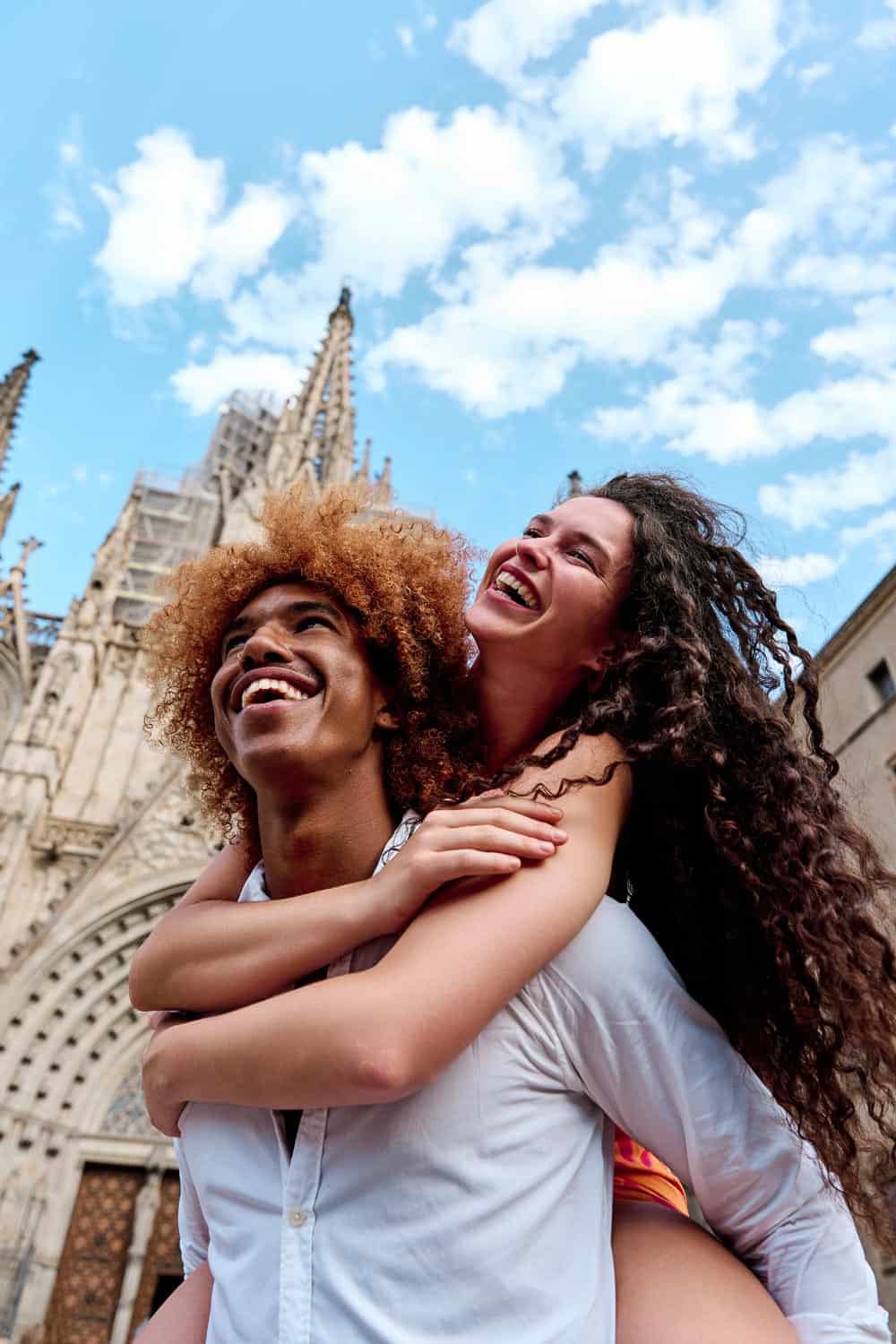 Two people smiling as one gives the other a piggyback ride in front of a Gothic-style cathedral in Spain under a blue sky with clouds.