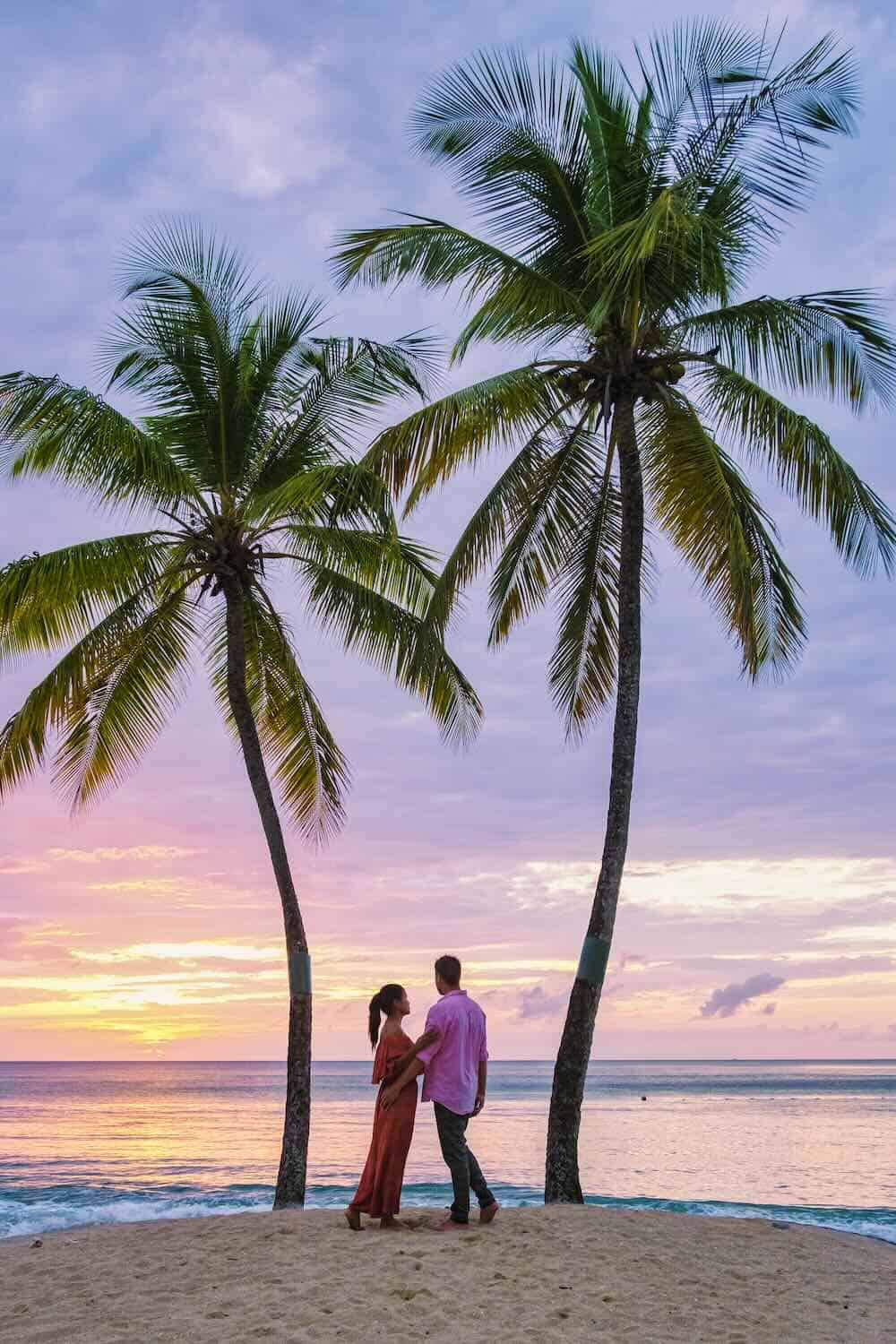 A couple stands on a St. Lucia beach at sunset, holding hands under two tall palm trees, with the ocean and colorful sky in the background.