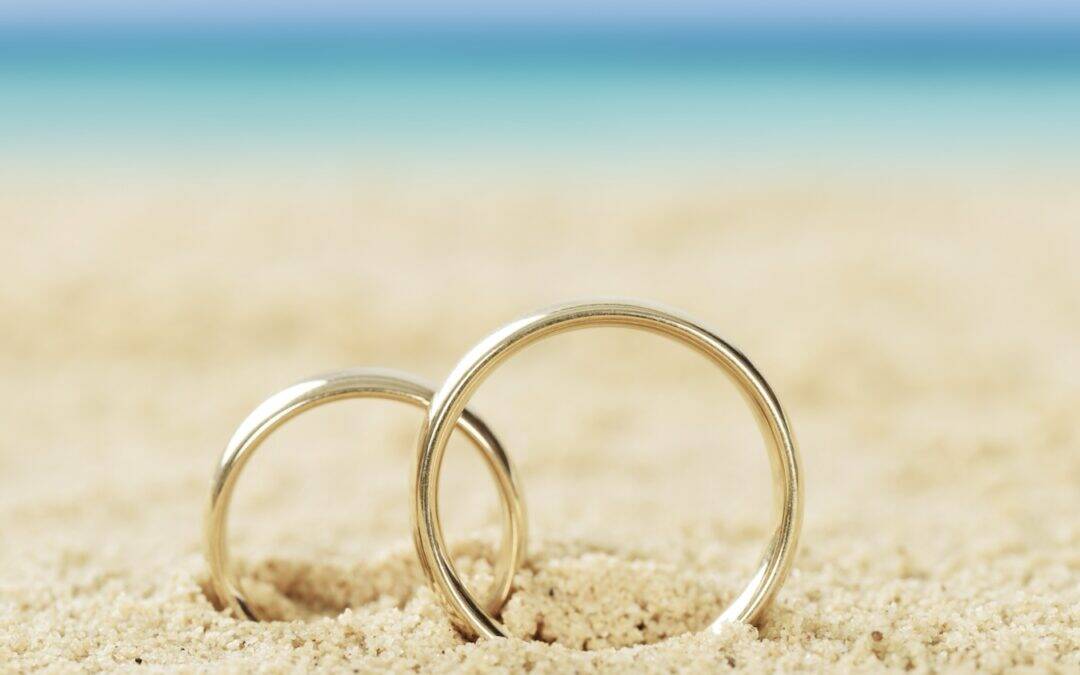 Two gold wedding rings are partially buried in sand on a beach, with the sea and sky blurred in the background.